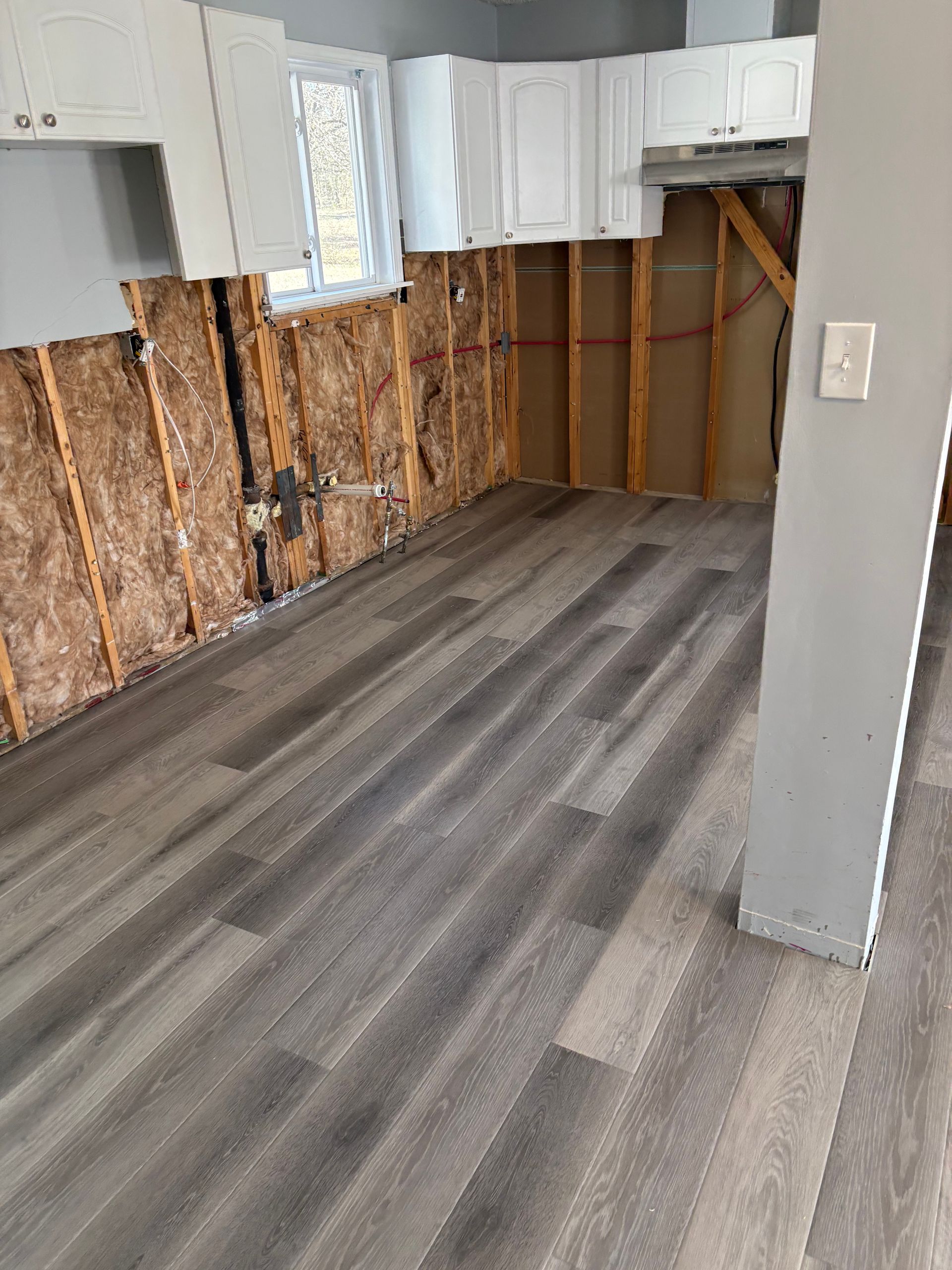 Partially renovated kitchen with exposed wall studs, white cabinets, and gray wood-look flooring
