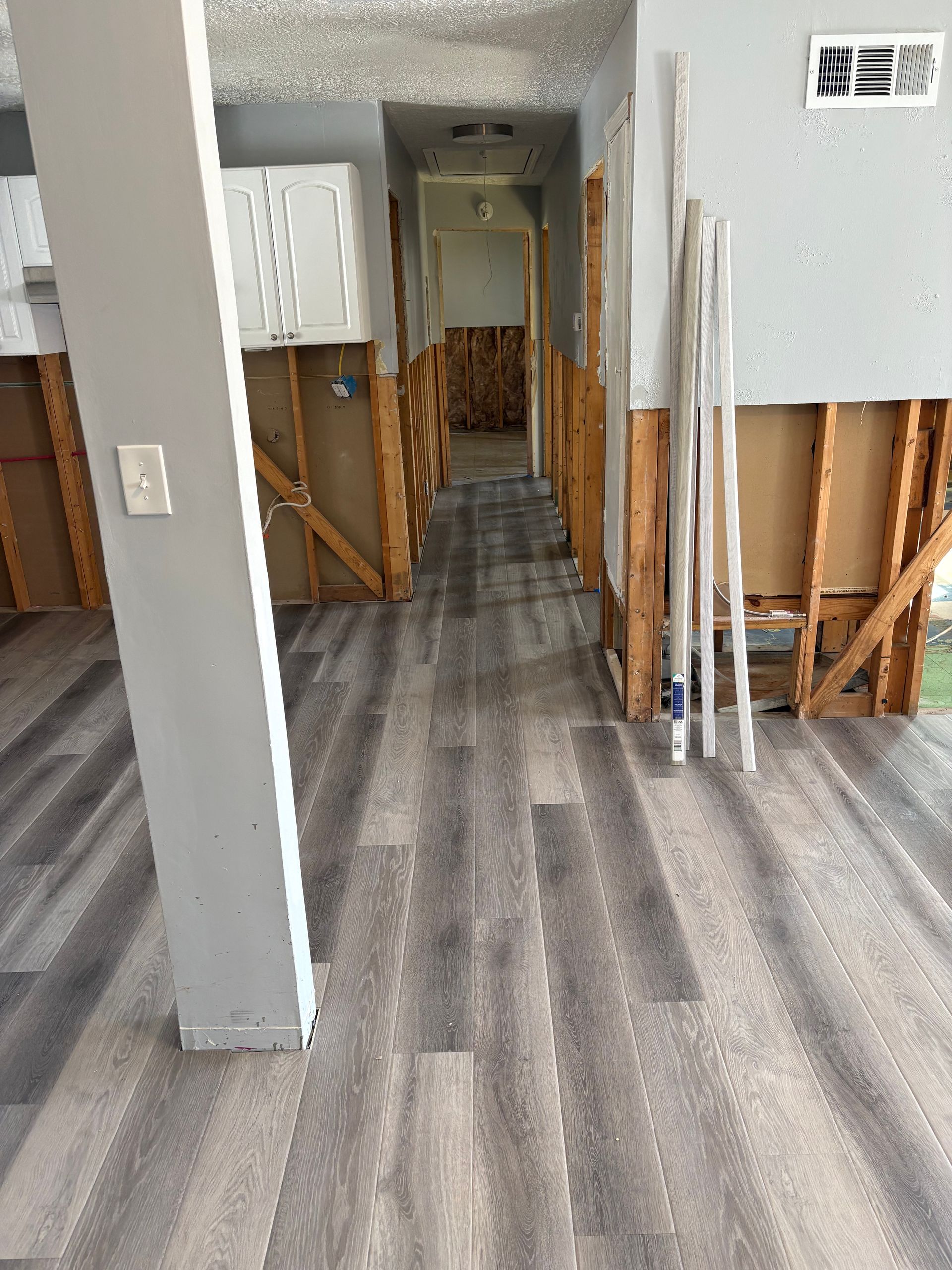Partially renovated hallway with exposed wall studs, gray flooring, and stacked trim against a white wall