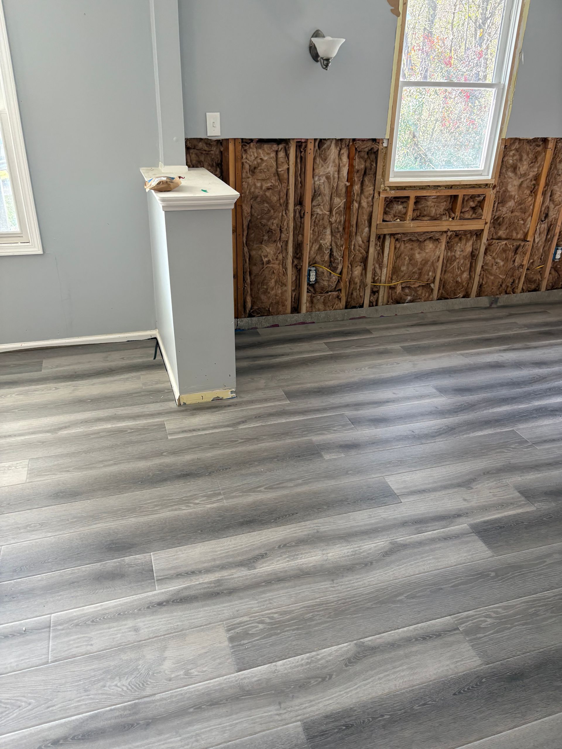 Empty room with gray wood flooring, exposed wall studs, and a small white pillar near a window.