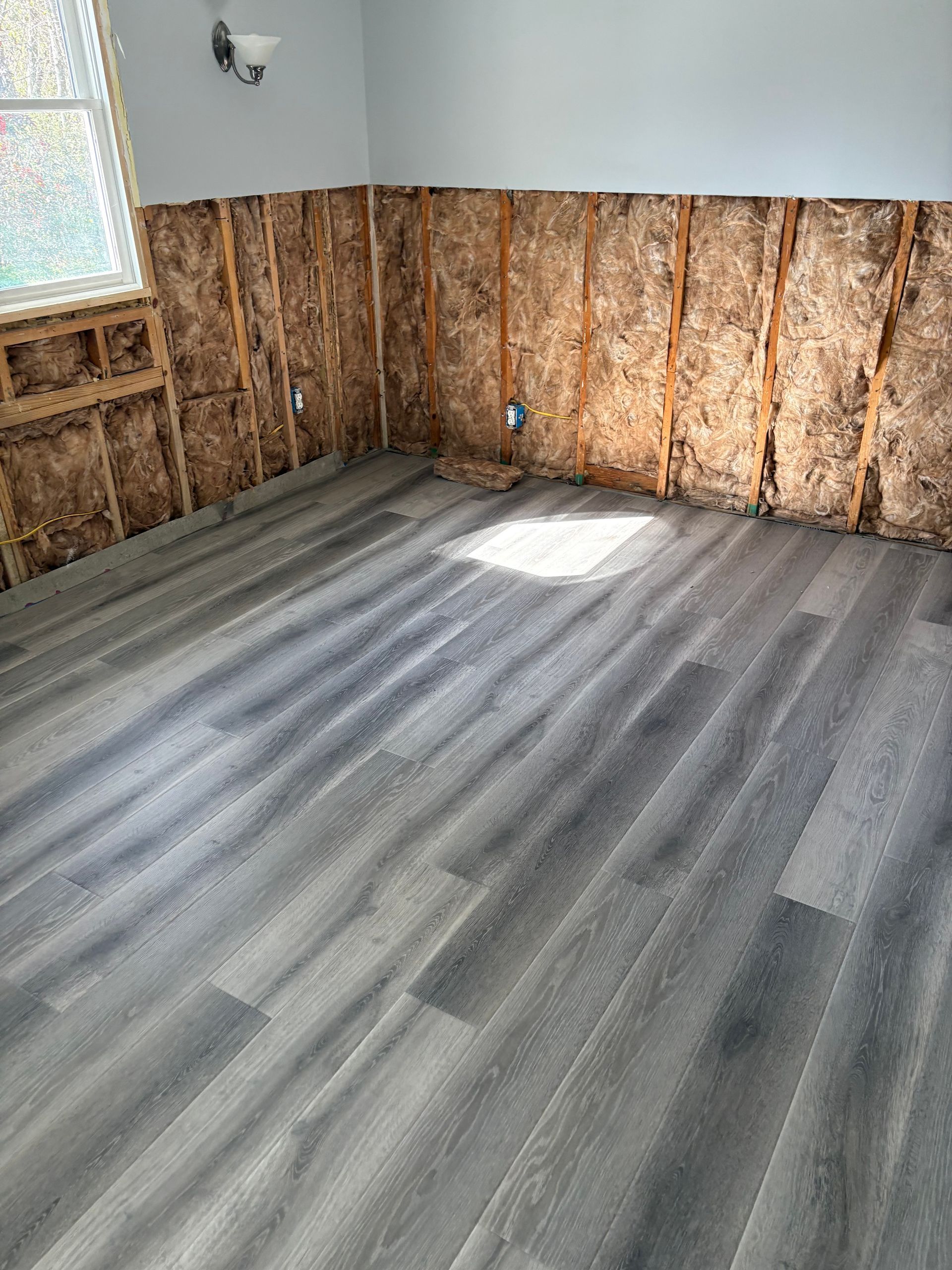 Empty room with exposed wall studs, gray vinyl plank floor, and sunlight near a window.