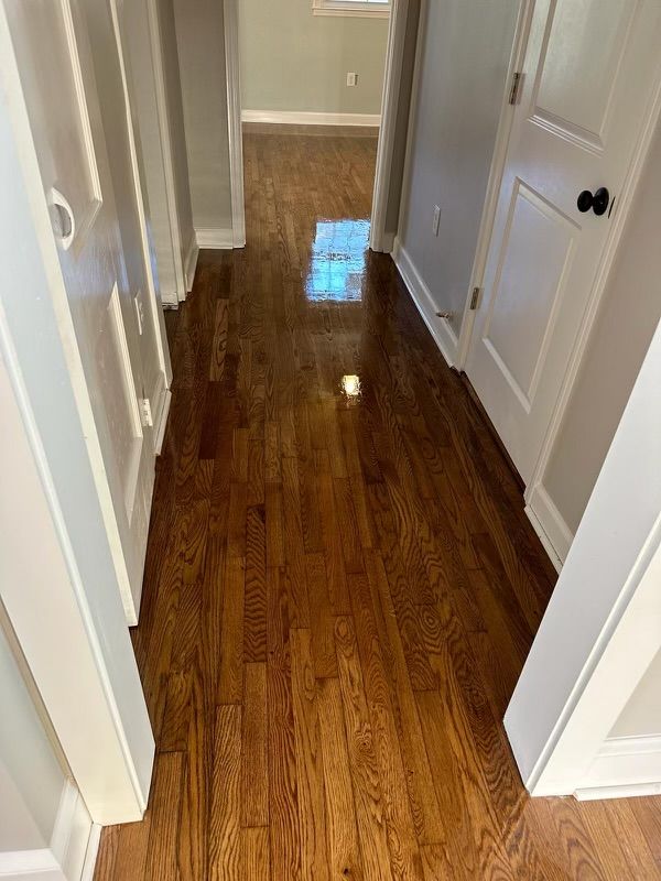 Narrow hallway with glossy hardwood floor, white doors, and tan walls leading to a windowed room.