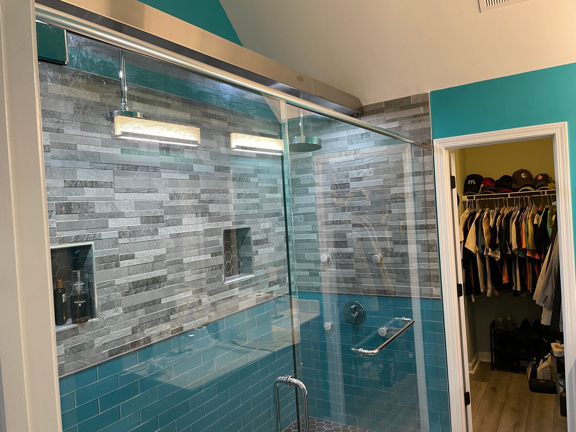 Modern bathroom with glass shower enclosure, gray tile walls, blue tub, and an open closet nearby