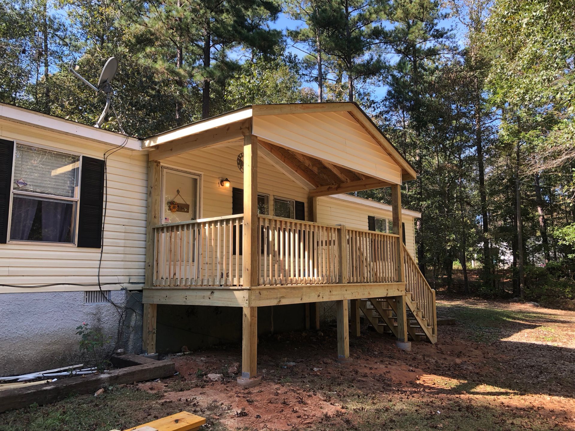 Small house porch with railing and steps in a wooded yard, sunlight filtering through pine trees