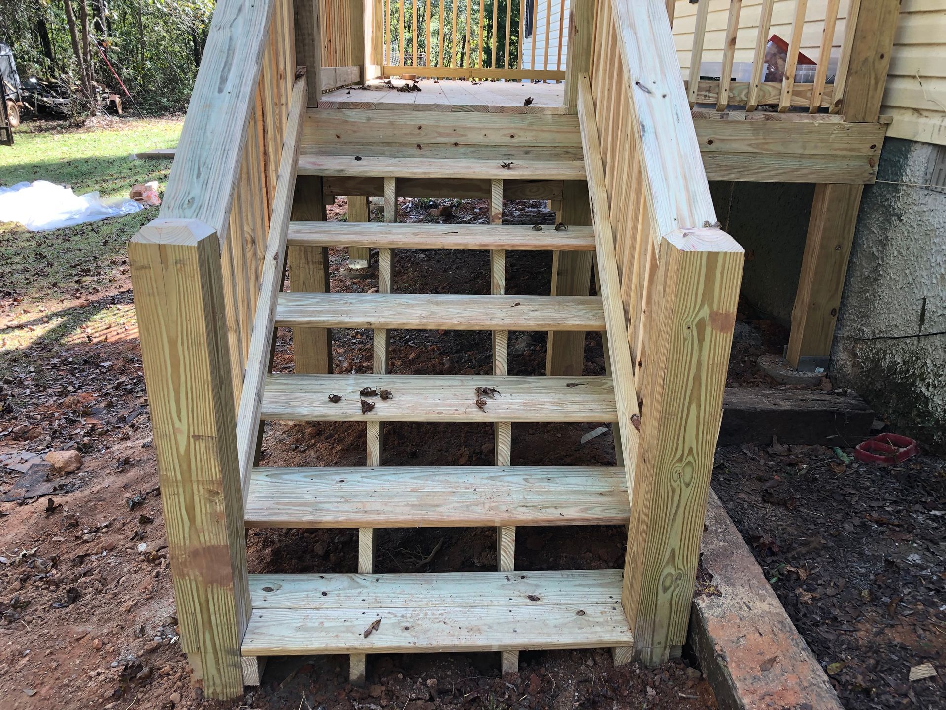 Wooden outdoor stairs with side railings leading up to a raised deck on a dirt and leaf-covered yard