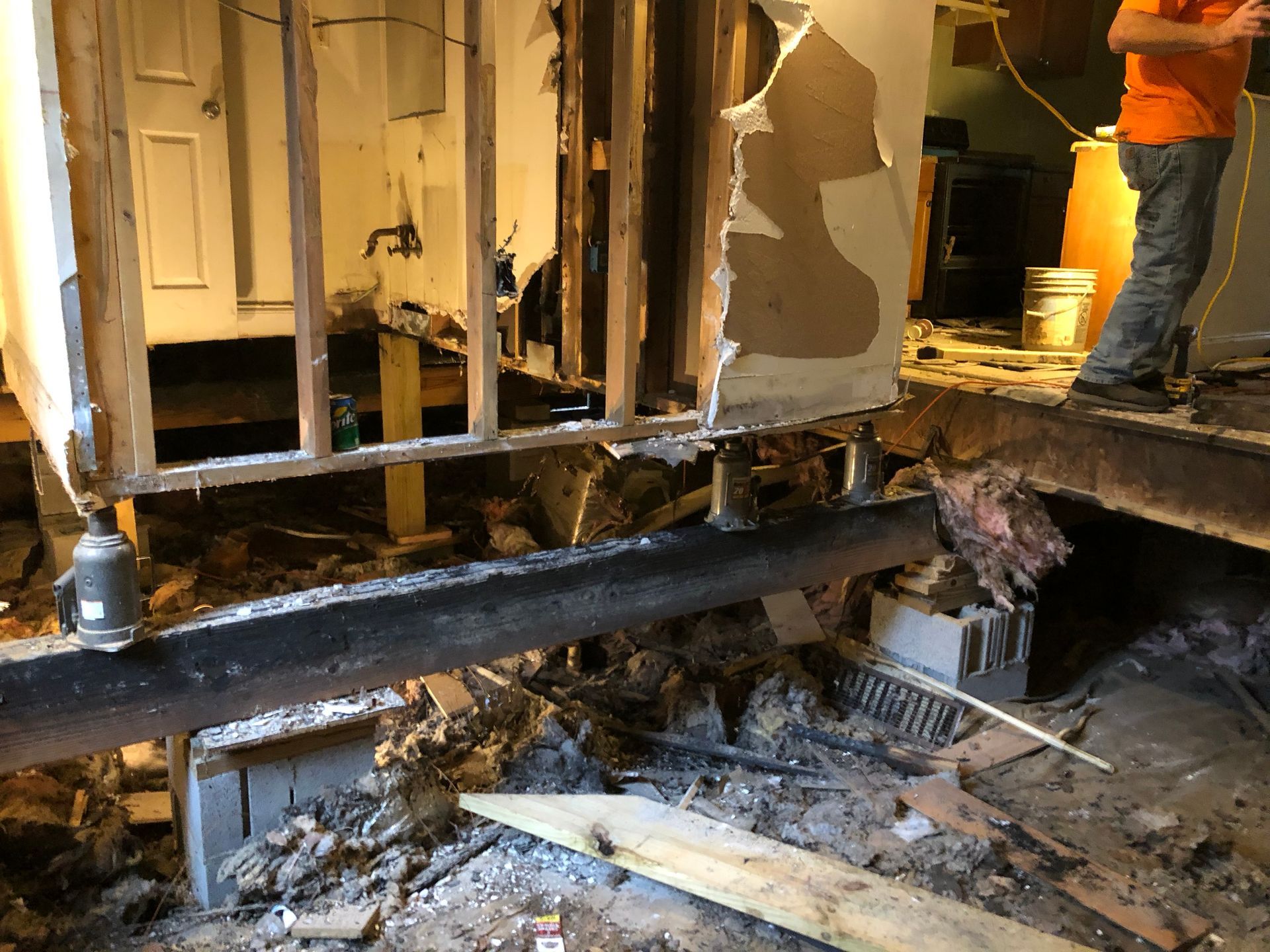 Damaged room with exposed floor joists, debris, and a person standing on a raised platform.