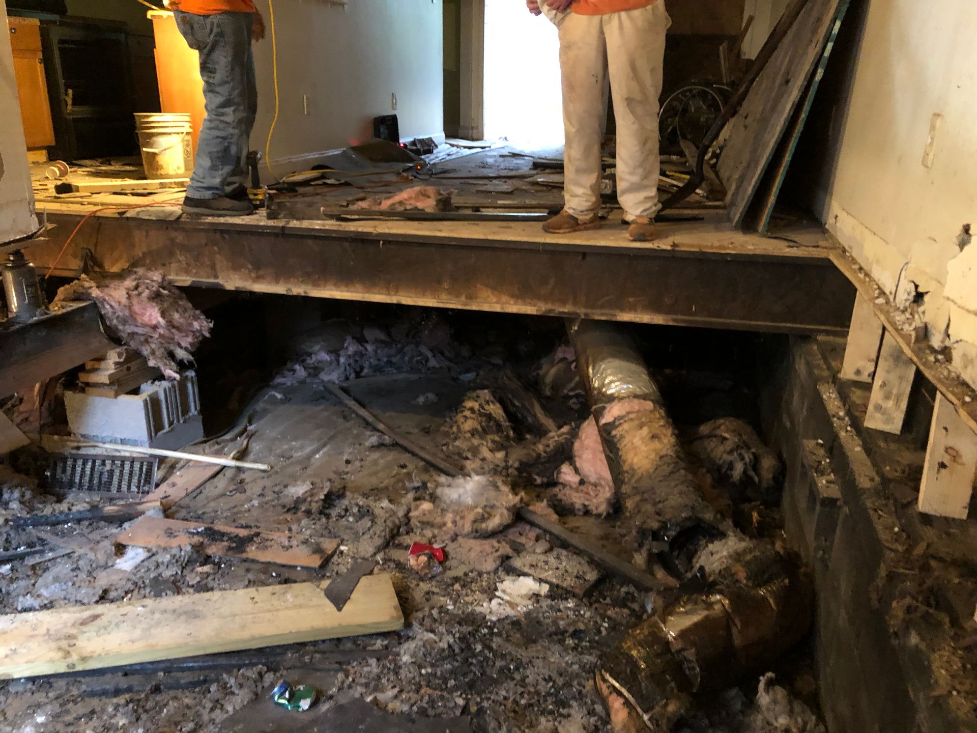 Collapsed floor exposing debris and pipes, with two people standing on the damaged edge indoors.