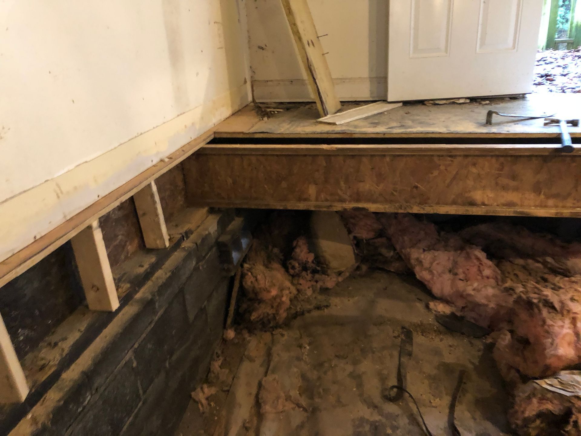 Exposed crawlspace under a floor, with dirt, support beams, and a partially open hatch inside a room