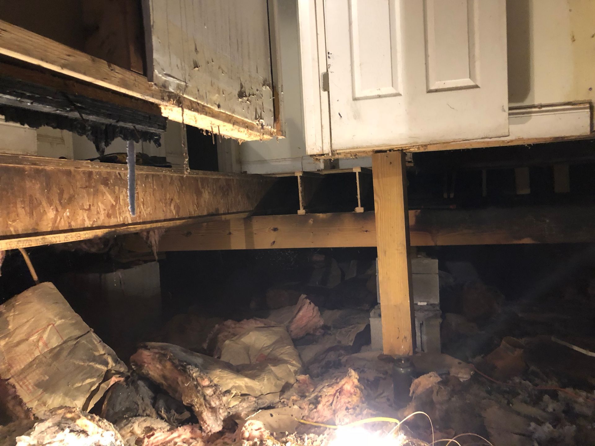Damaged kitchen floor with exposed joists, debris, and white cabinets above.