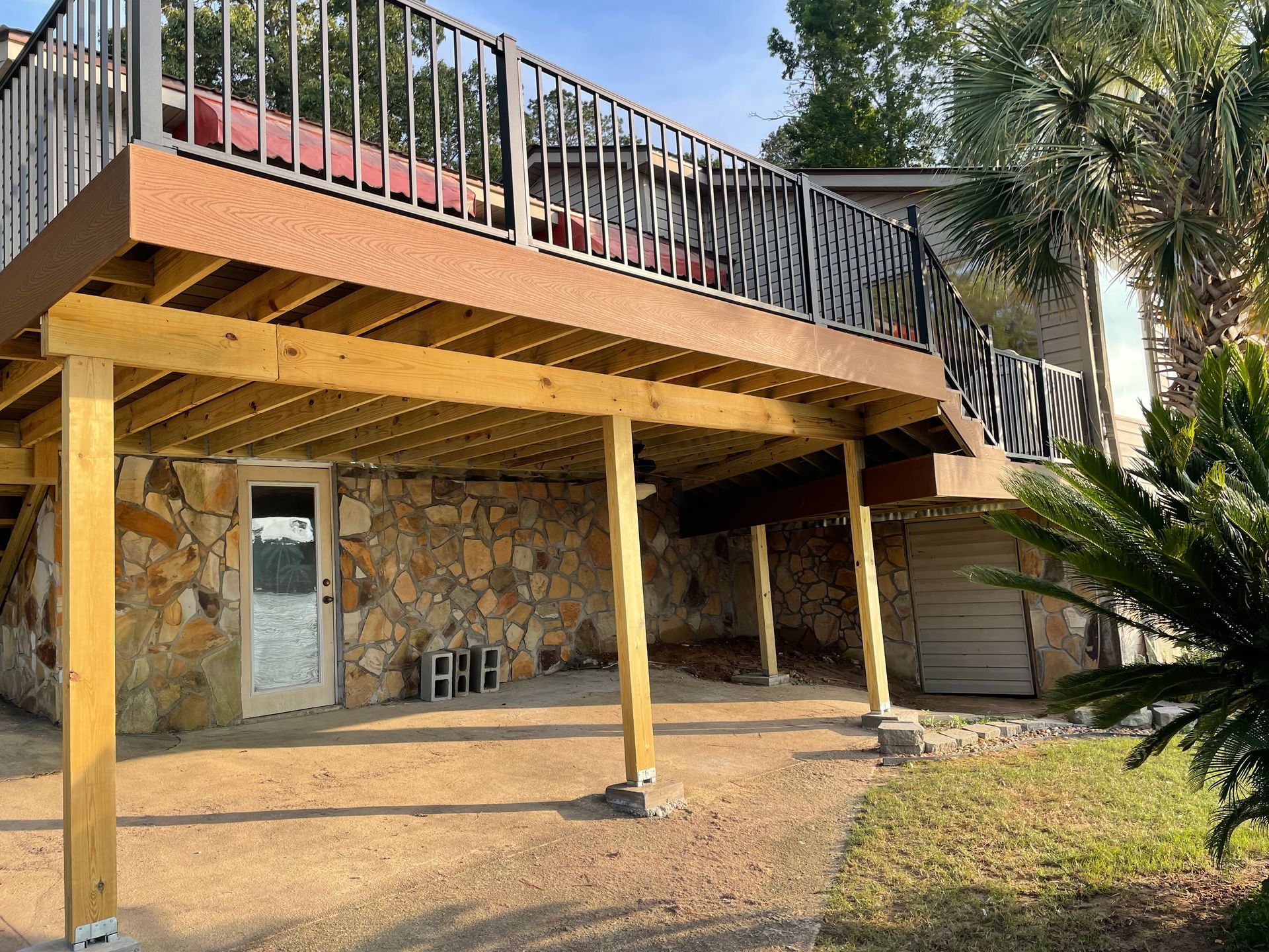 Wooden deck on stilts attached to a stone house, with a metal railing and stairs in a yard.