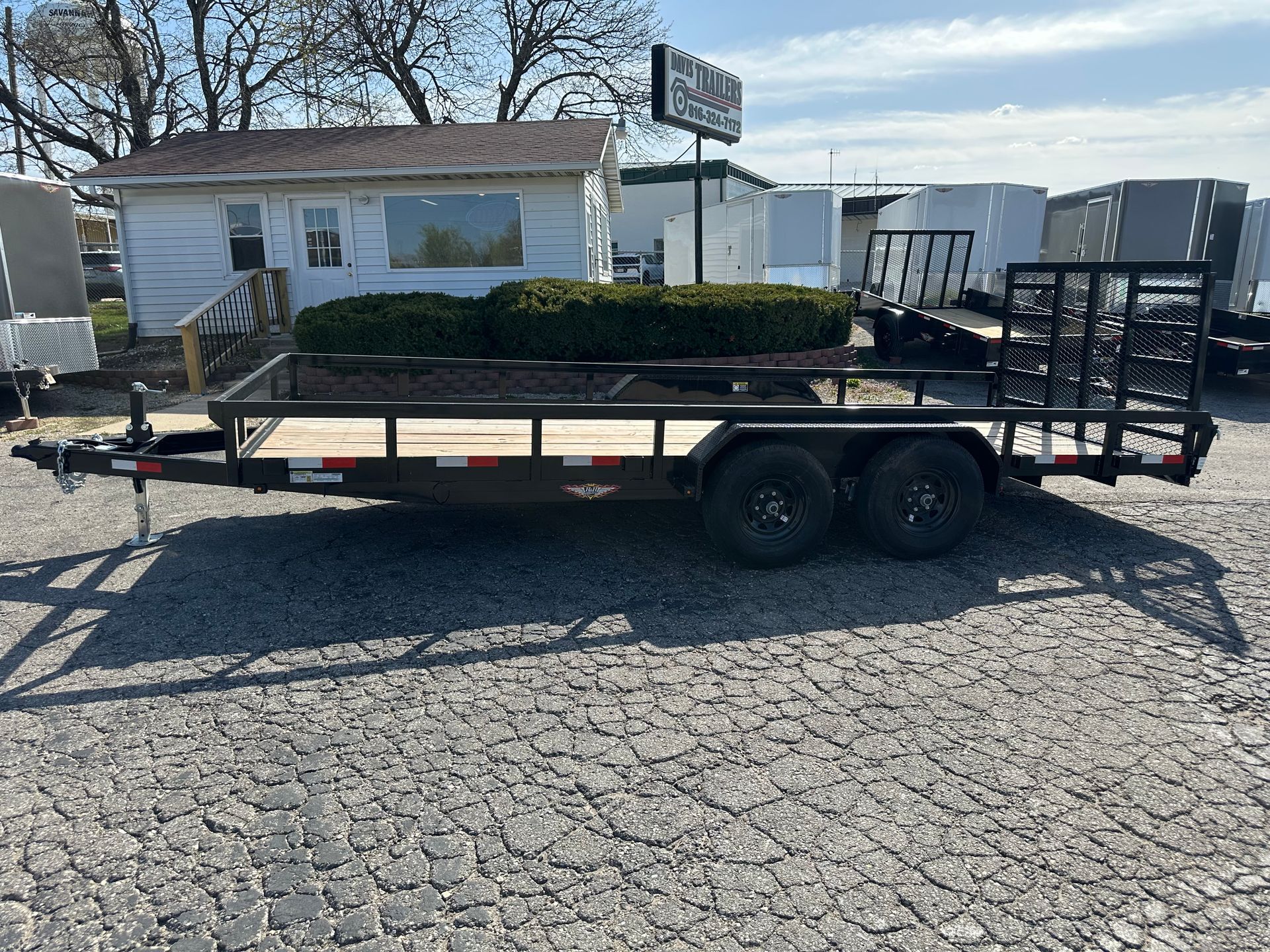 Black utility trailer with dual wheels, parked on gravel. A small building is in the background.