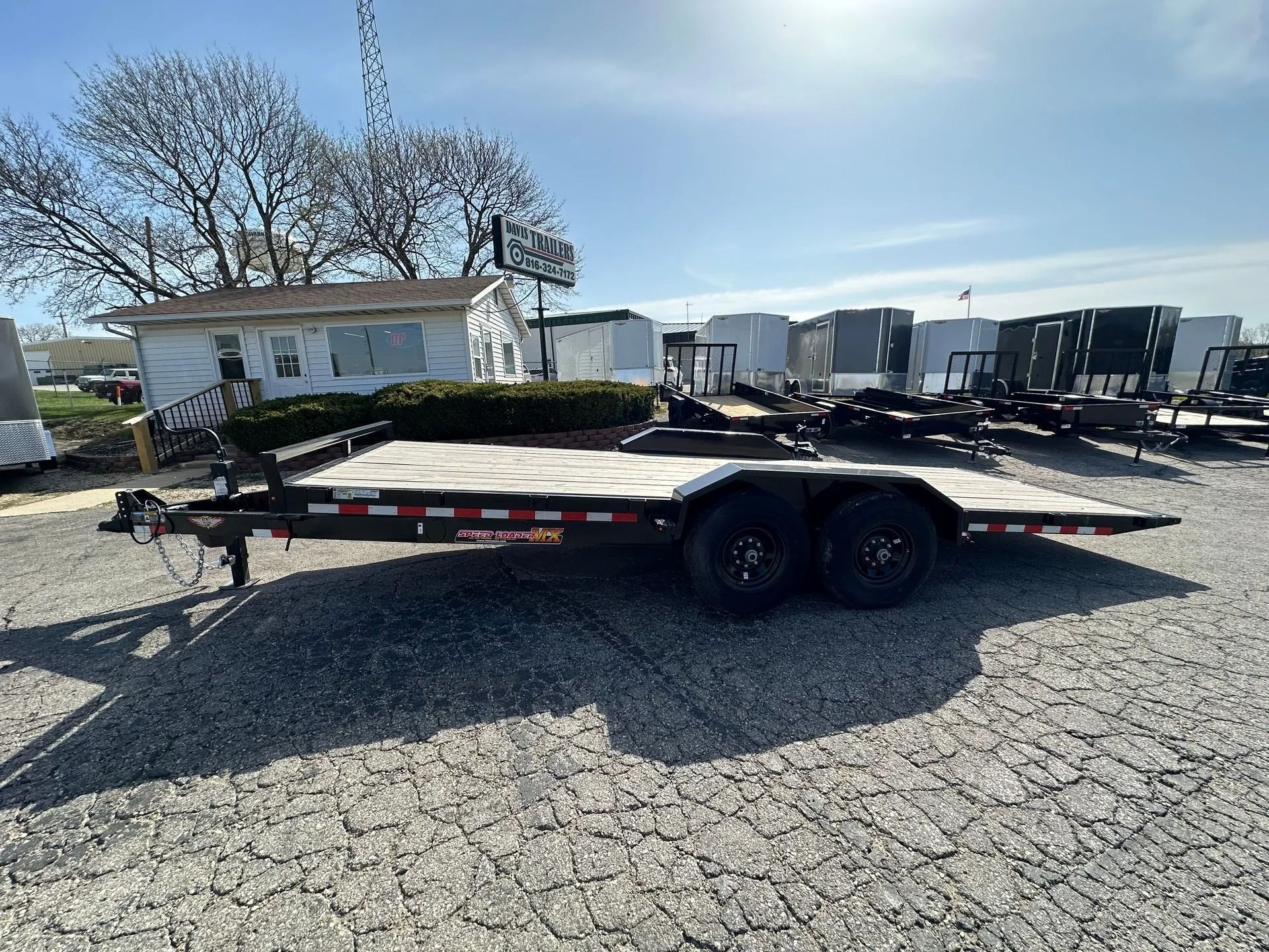 Black dual-axle flatbed trailer parked on cracked asphalt in front of a small building on a sunny day.
