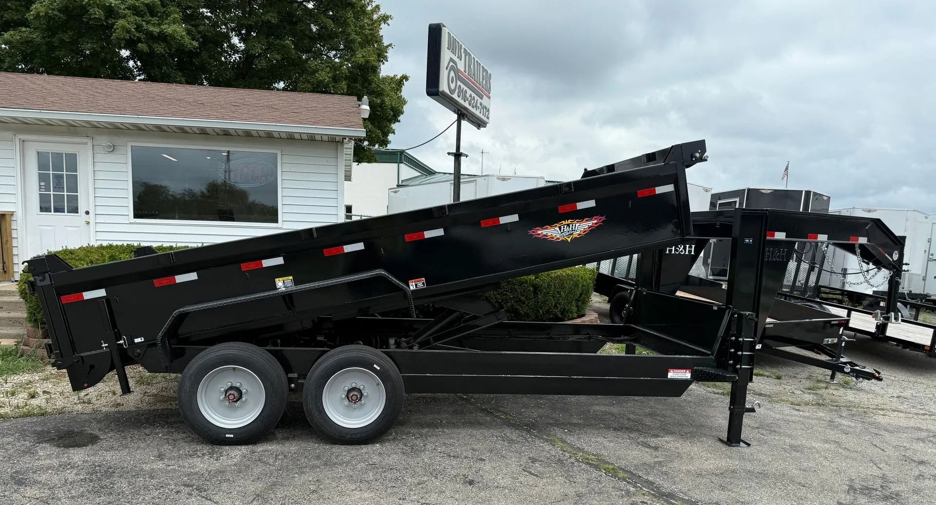 Black dump trailer with two wheels, tilted up, parked outside a building.