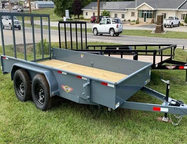 Gray utility trailer with black tires on grass, road and buildings in background.