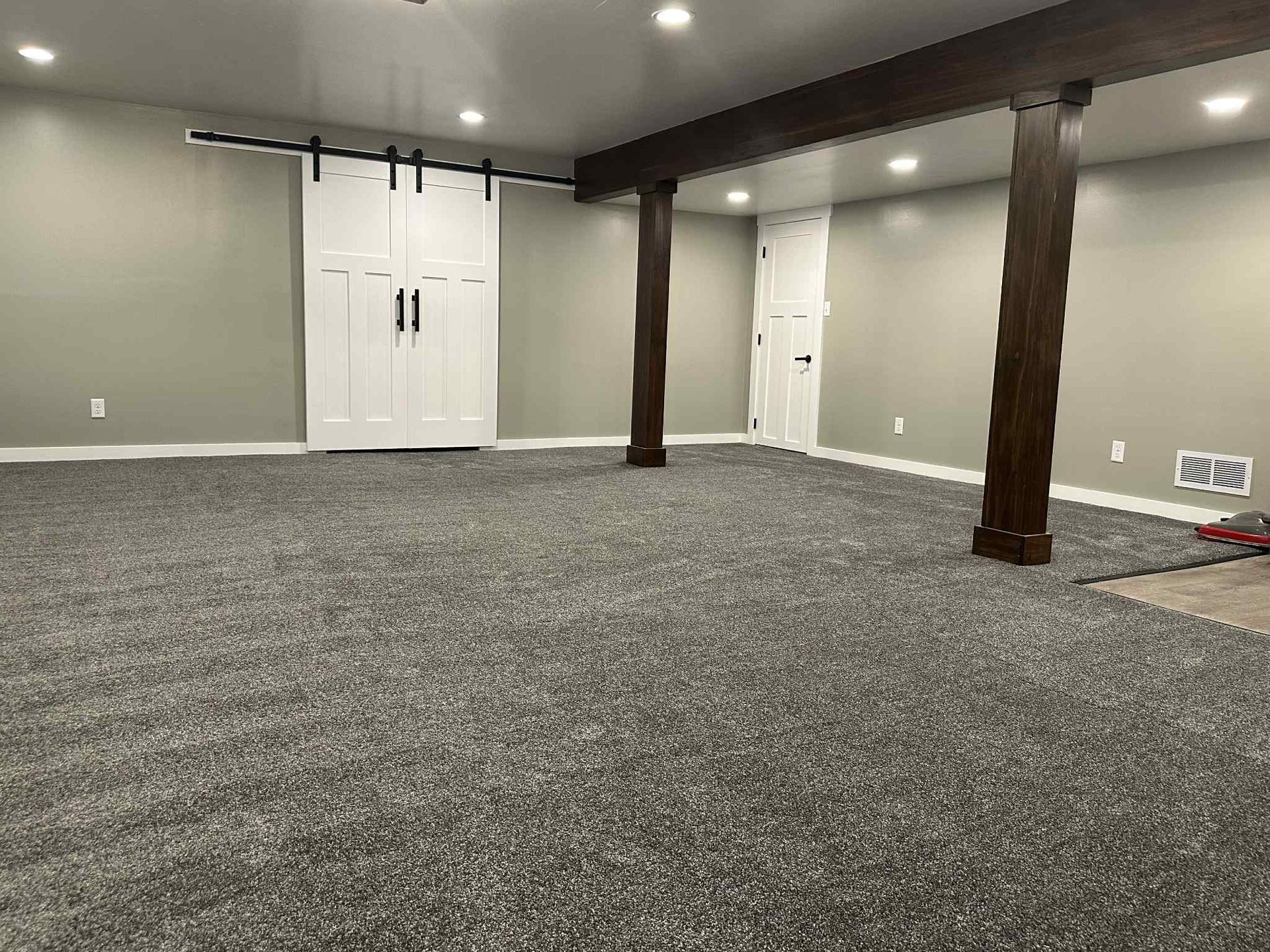 Gray carpeted basement with white sliding doors and dark wood support beams.