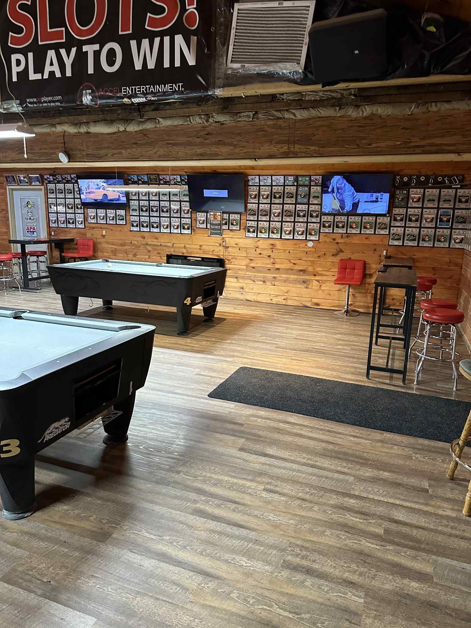 Pool hall with two pool tables, TVs, red bar stools, and wooden walls.
