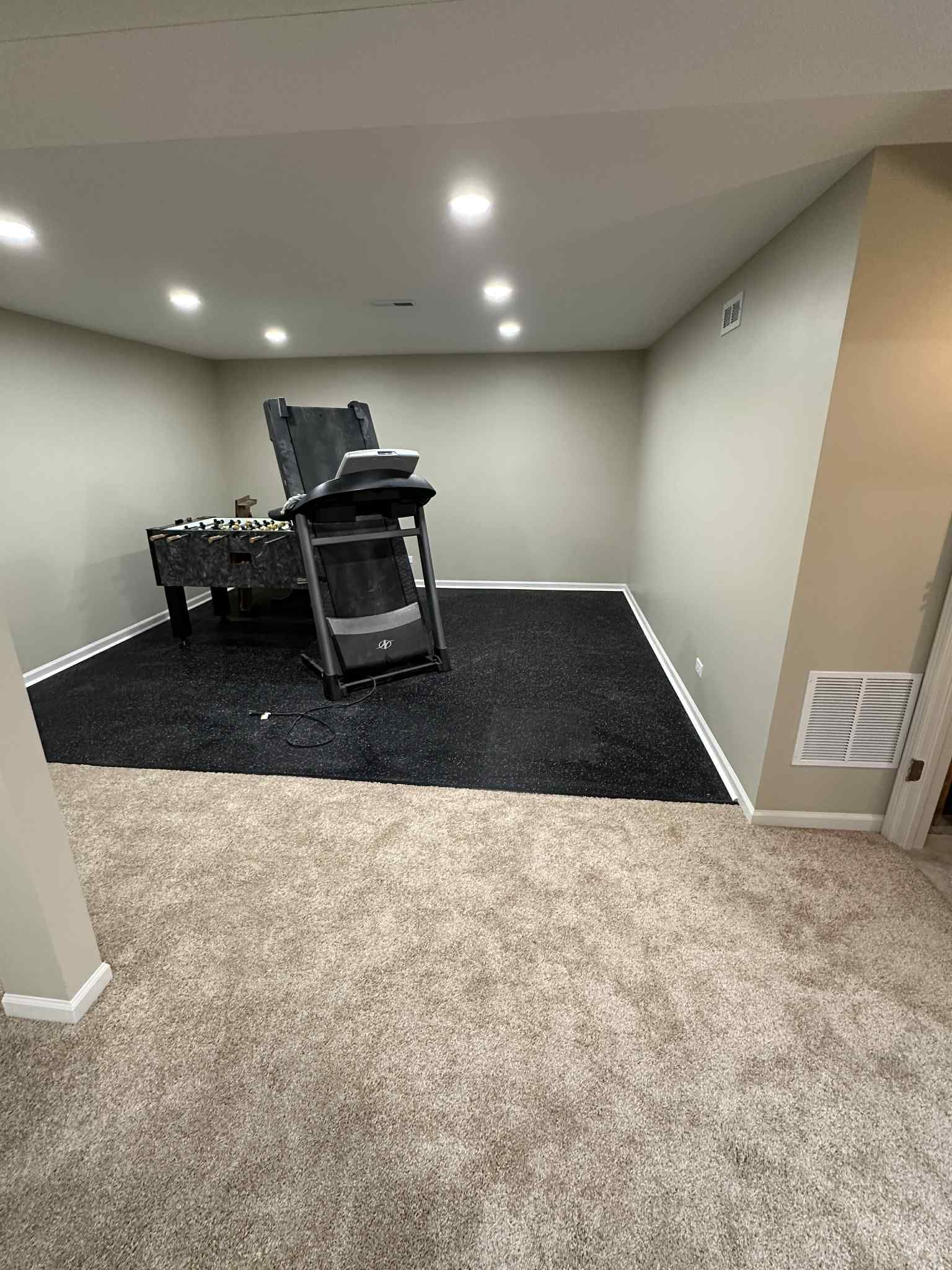 A treadmill on black rubber flooring in a basement gym with beige carpet.