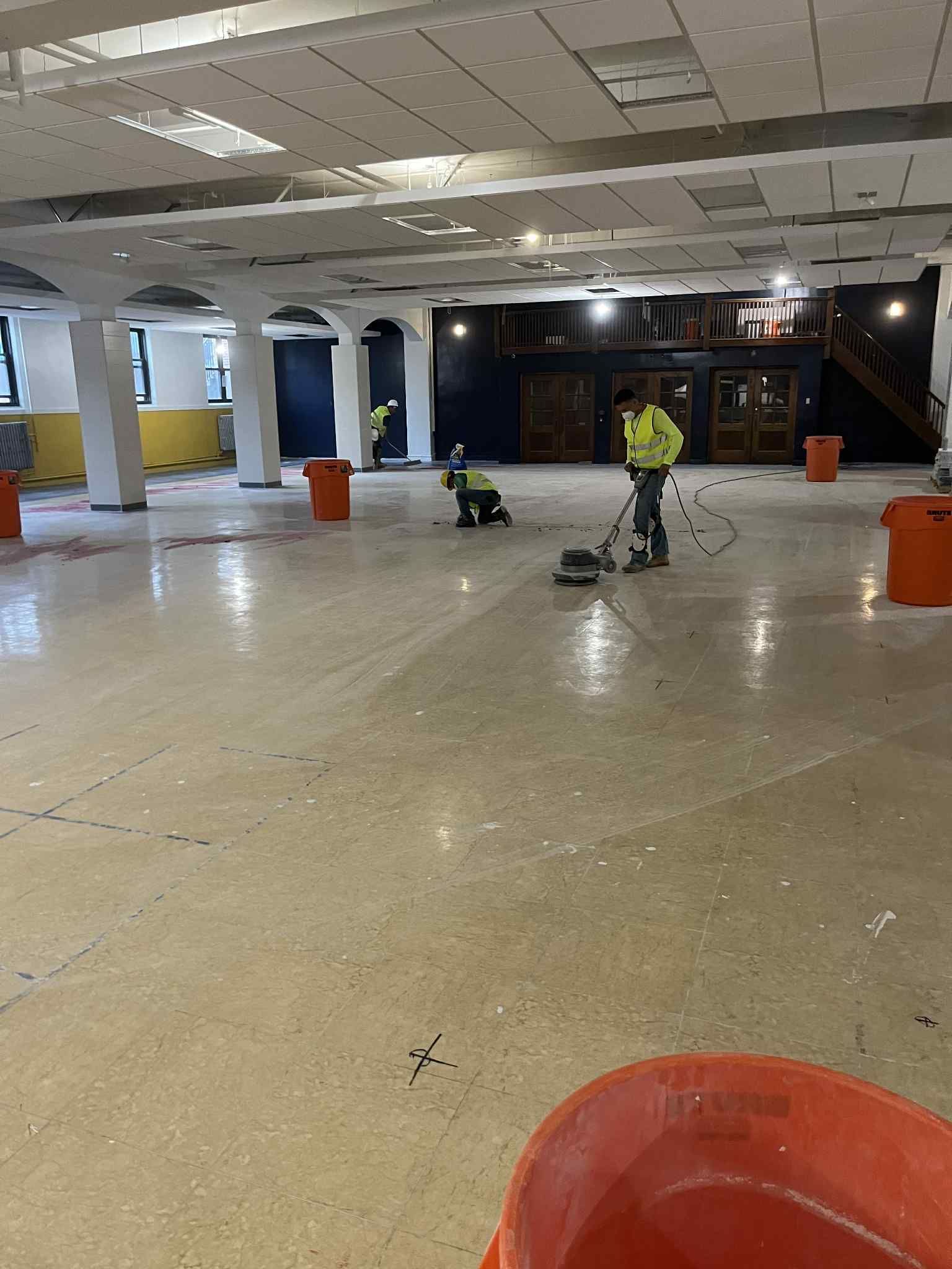 Workers in neon vests grinding a large, open floor. Orange buckets are scattered around.