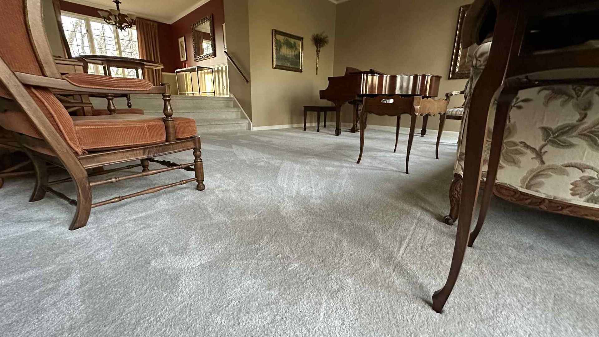 An antique chair and table sit on light blue carpet, with a piano and staircase in the background.