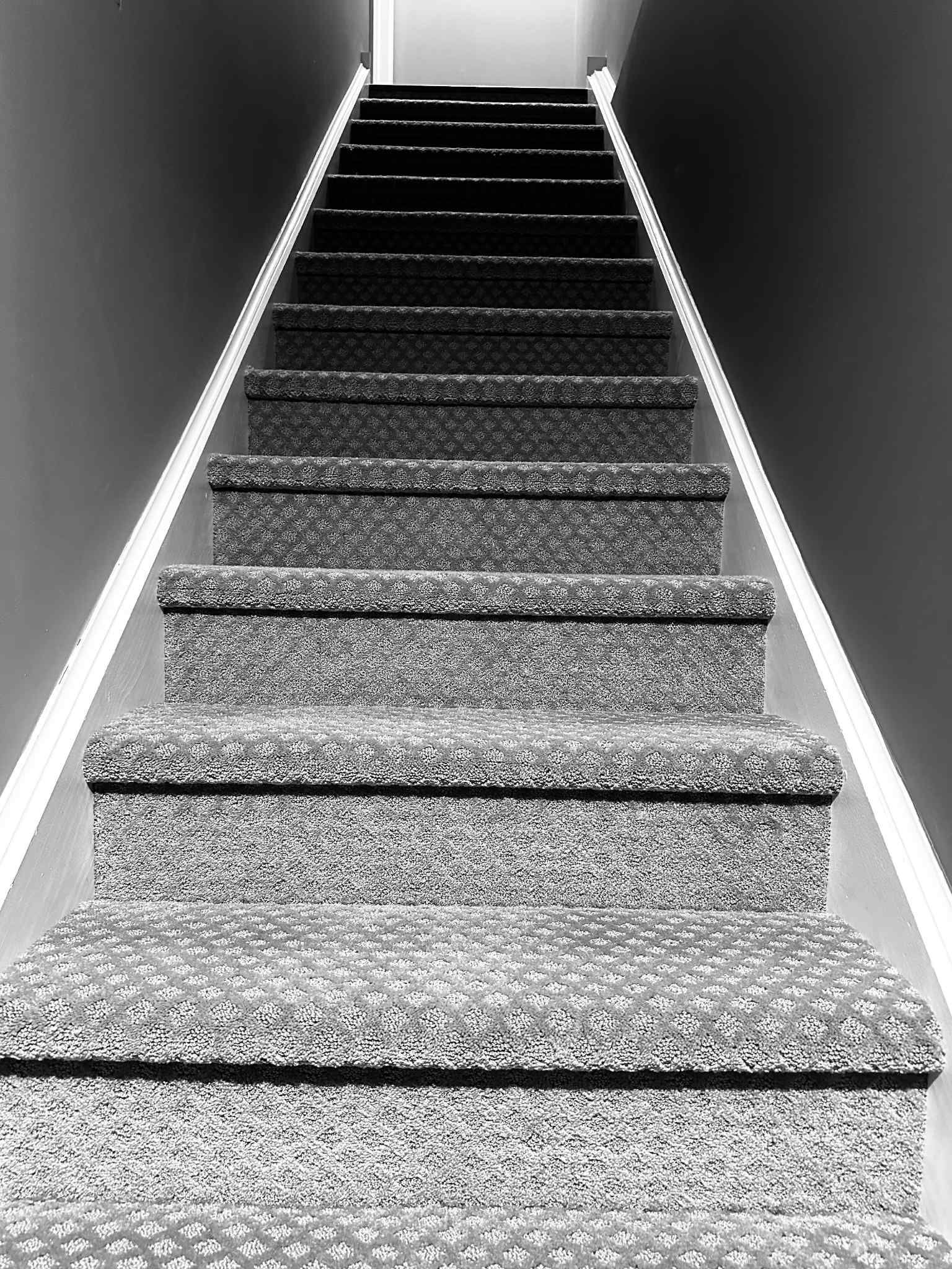 Black and white image of carpeted staircase leading upward toward a light at the top.