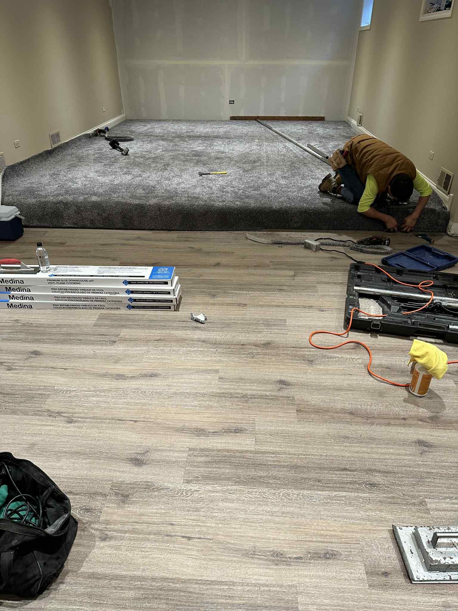 Person installing carpet in a room with wood-look flooring. Tools and materials are scattered around.