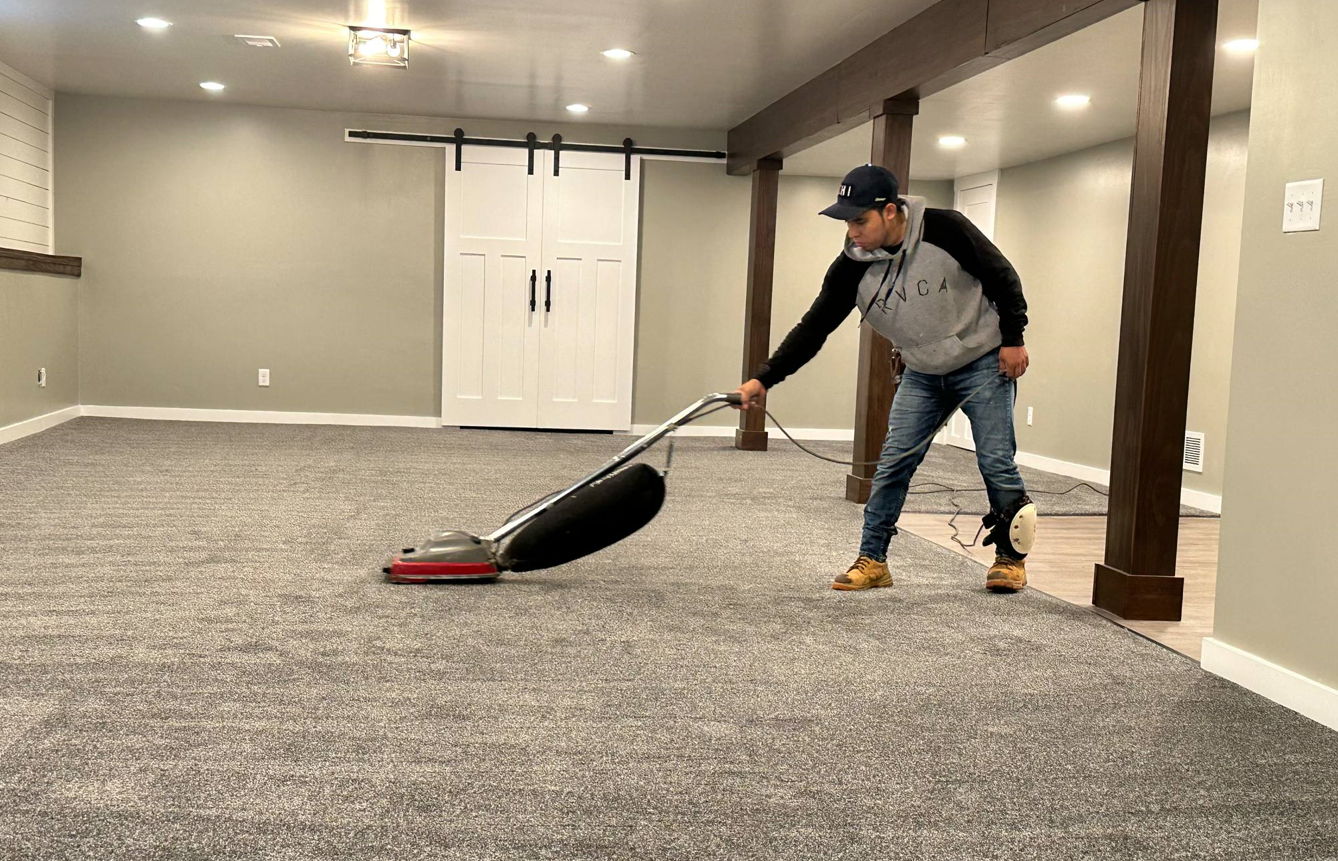 A person vacuuming a gray carpeted room with wooden support beams and a white sliding door.