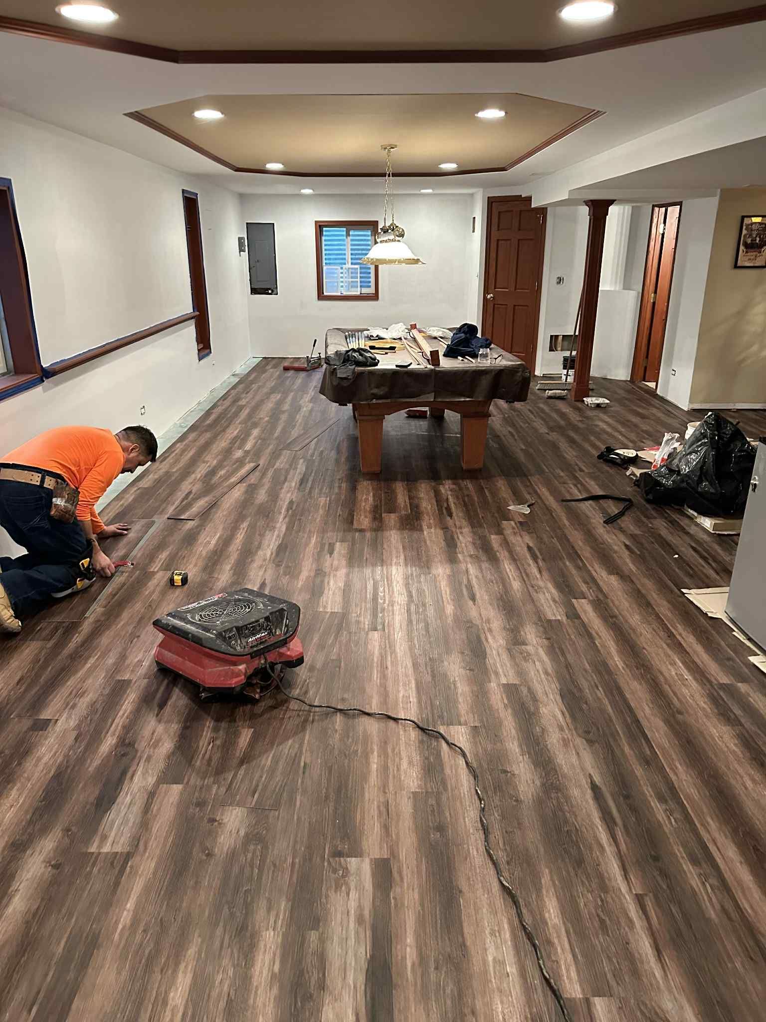 Man installing dark wood-look flooring in a basement with a pool table.
