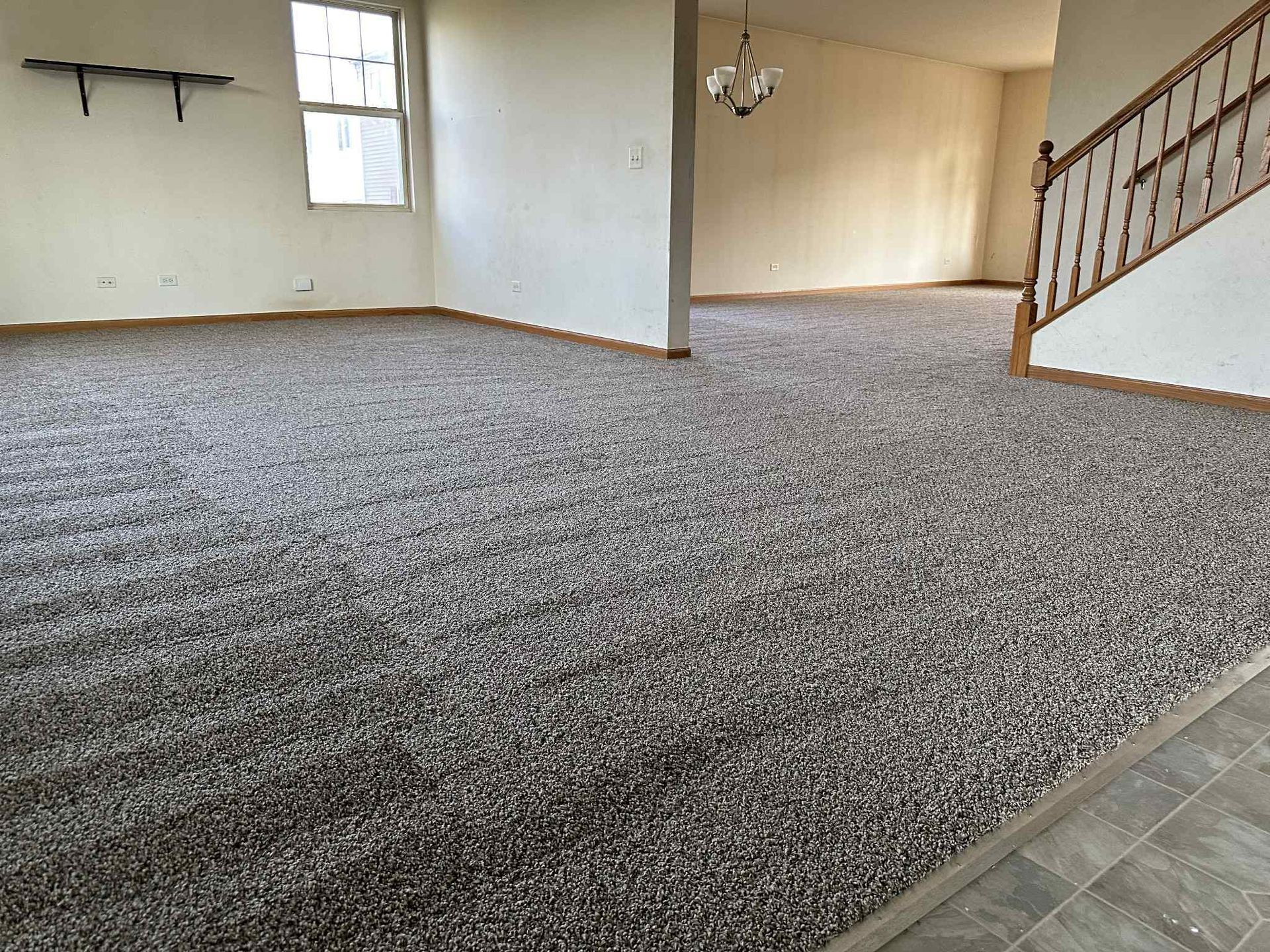 Spacious room with dark speckled carpet, beige walls, and a staircase. A chandelier hangs in the distance.