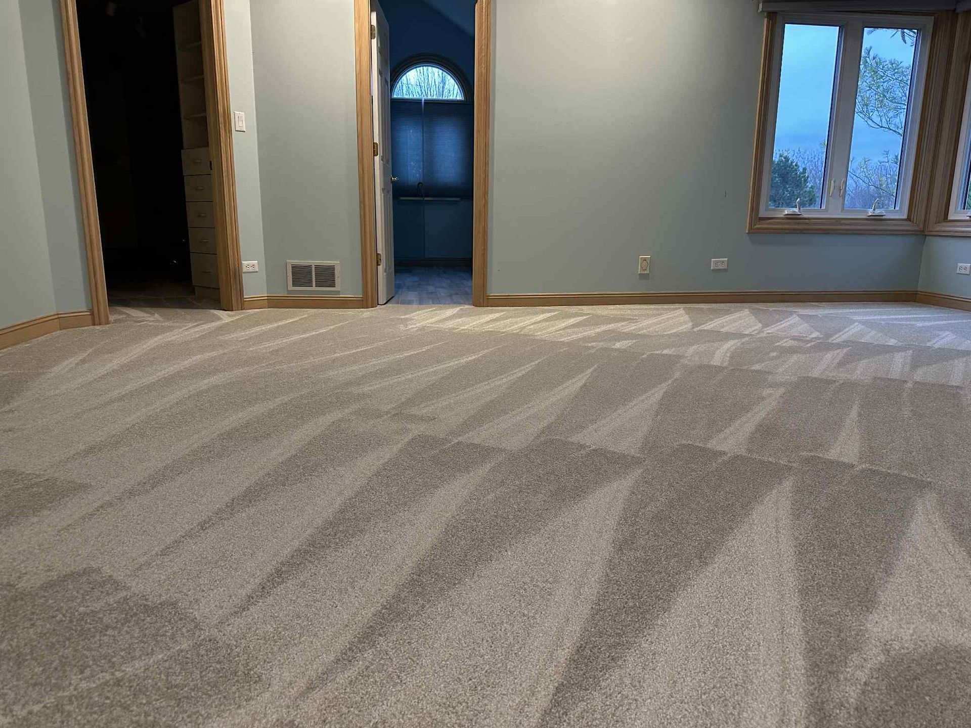 Room with gray patterned carpet, pale blue walls, wooden trim, and windows.