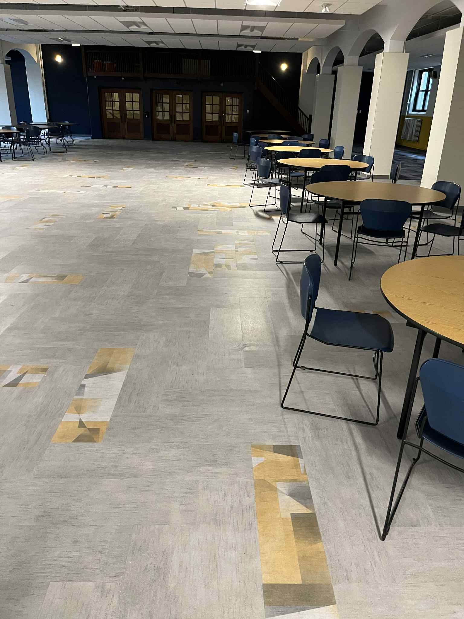Empty cafeteria with round tables and chairs; faded floor tiles; arched columns.