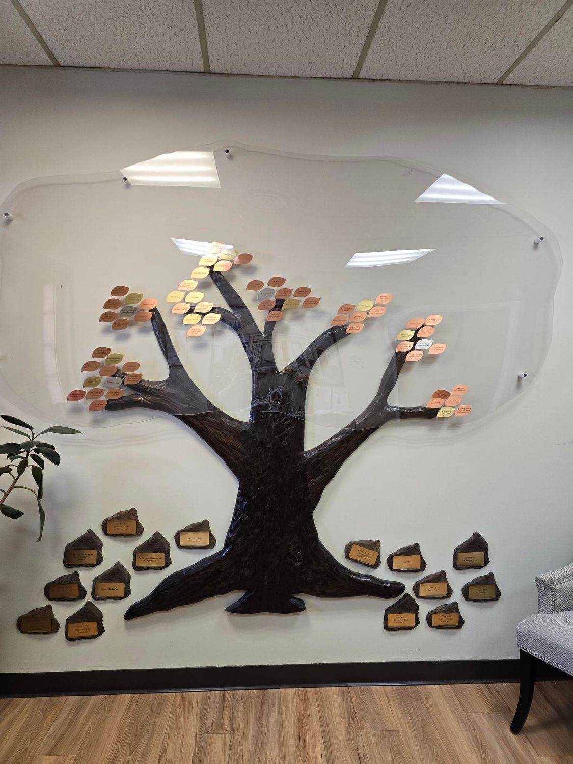 Wall-mounted tree sculpture with bronze leaves and plaques, under a clear acrylic shape.