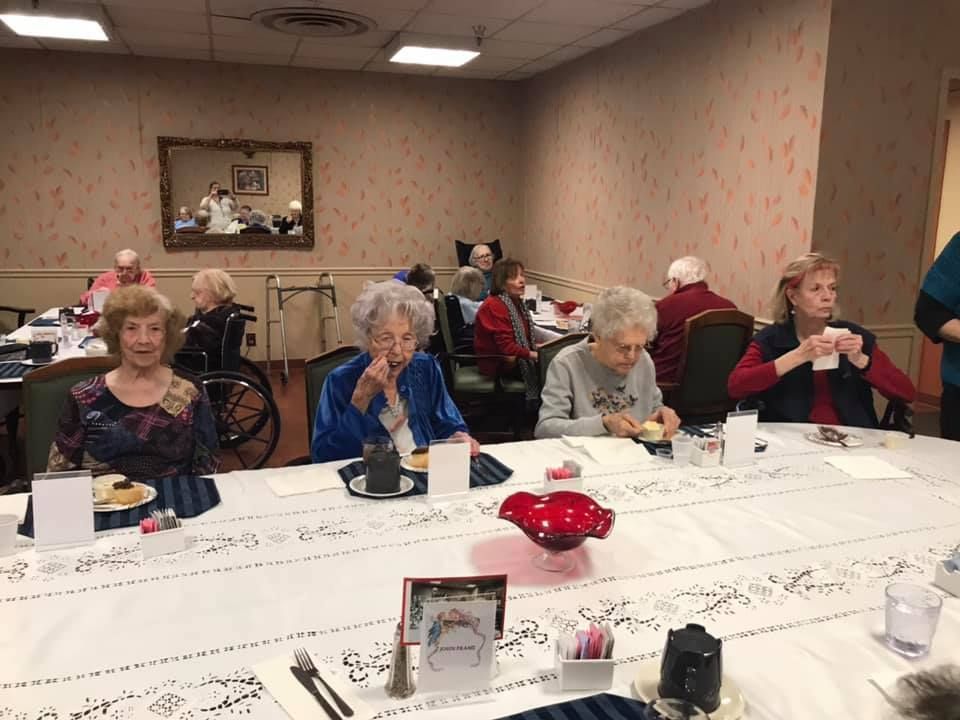 People seated at a long table, possibly in a dining room, some are eating or holding items, while others are interacting.