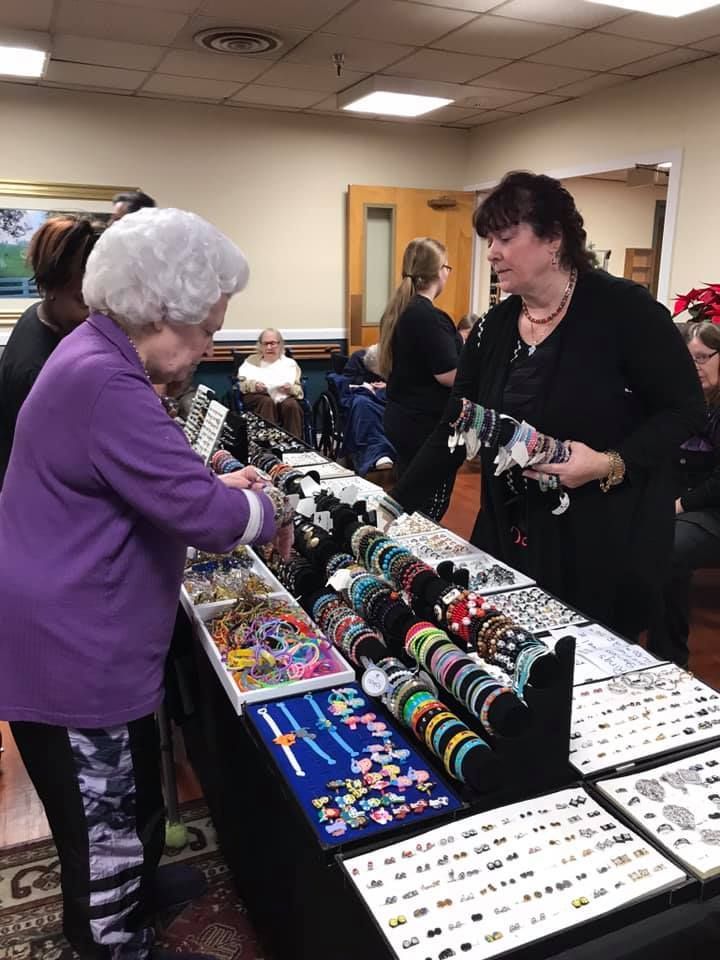 Woman examines bracelets at a table display; another woman assists. Indoor setting with additional shoppers.