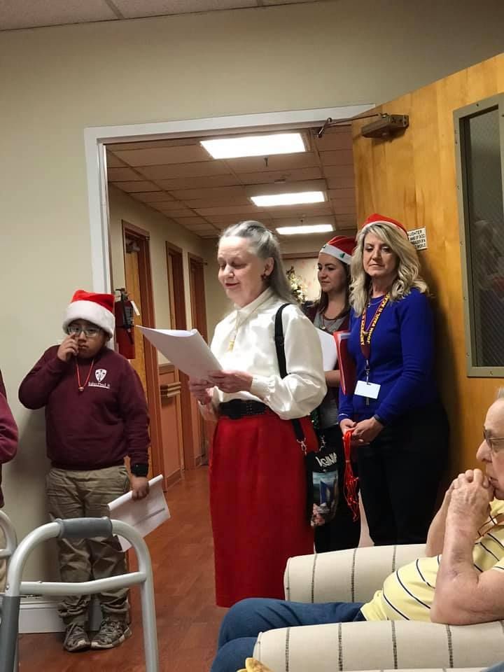 Group singing carols in a hallway. Person in red skirt reads from sheet, others wear Santa hats.