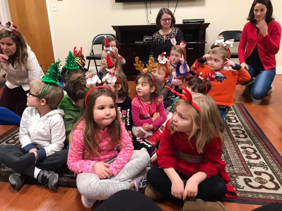 Children wearing holiday headbands sit on floor, looking at teacher. Adults in background.