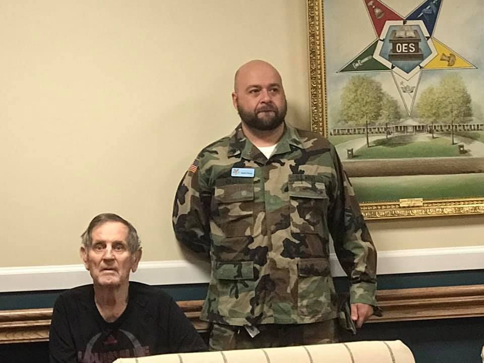 Man in camo uniform and older man seated, in front of a painting with a star.