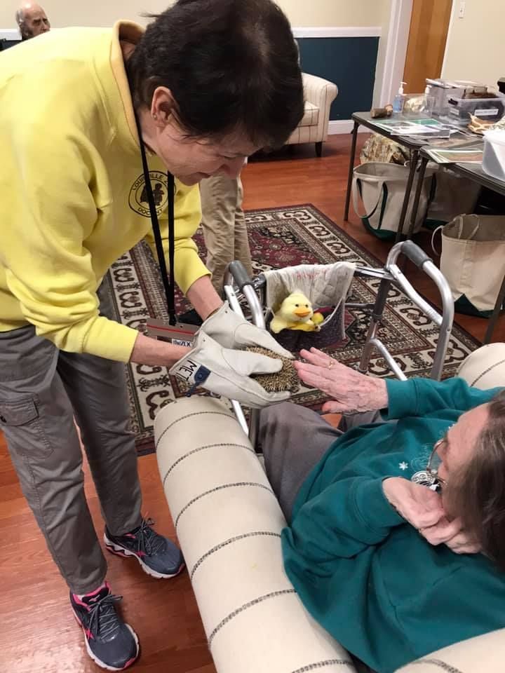 Woman in yellow shirt helps another person seated in a chair, with a walker in the background.