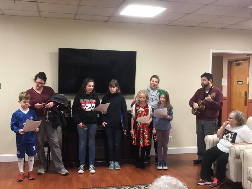 Children singing carols, holding papers, with adults, one playing ukulele, in a room.