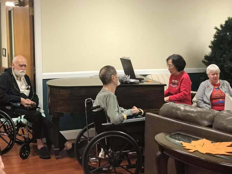 People in wheelchairs around a piano, one playing a laptop. Indoor setting.