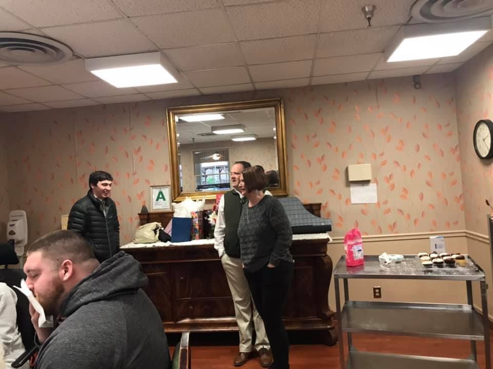 People in a room with a mirror and a cabinet. Two people stand near a cabinet talking. One man smiles.