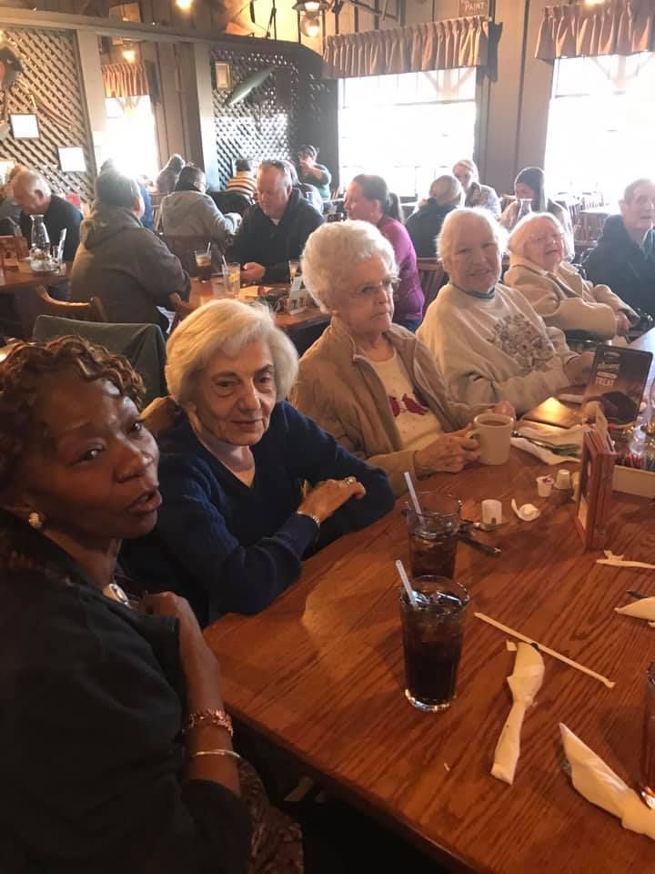People seated at a table in a restaurant; some looking at the camera, others in conversation. Dark wooden table, natural light.