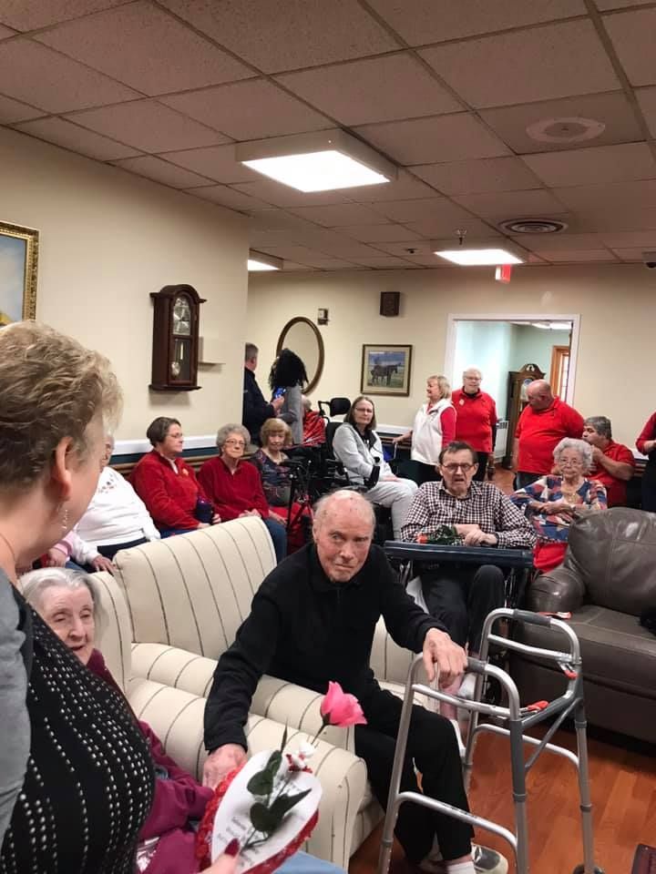 Seniors gather in a room, some seated on sofas. One man uses a walker. Others wear red shirts.