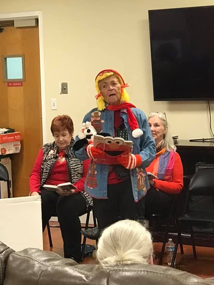 Woman in costume reading, wearing scarf, holding book. Two others seated behind, watching.
