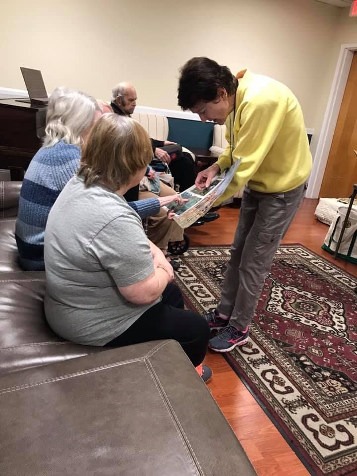 Group of people looking at something, one person is pointing. Indoors on a rug, with a piano in the background.