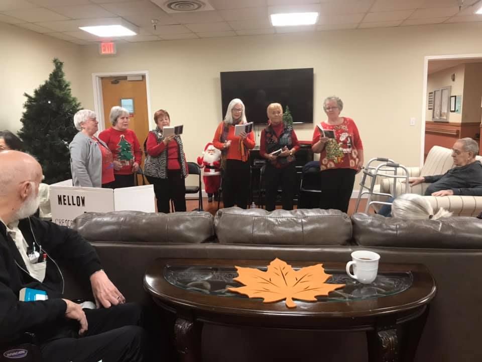 Group of people singing, some holding songbooks, in front of a seated audience; room decorated for holidays.