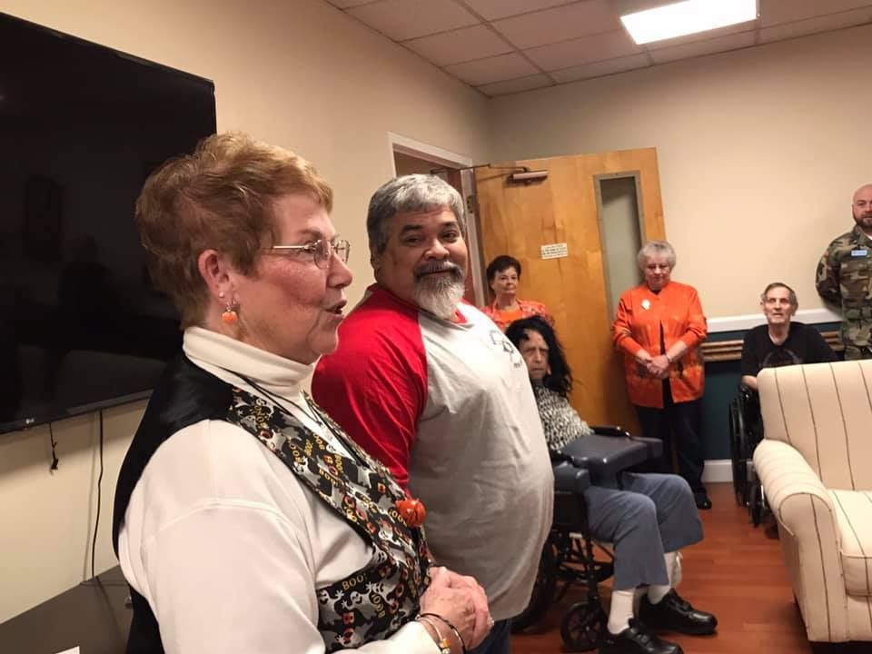 Group of people indoors, some standing and speaking, others seated.  One person is in a wheelchair.  Smiling and listening.