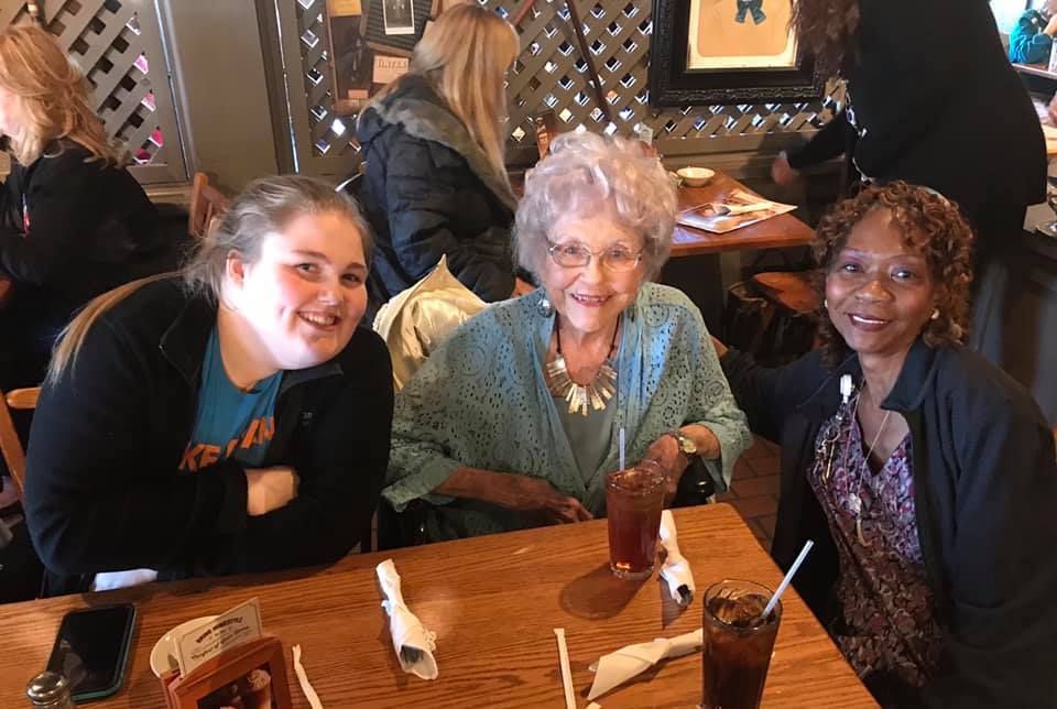 Three people smile at a table in a restaurant. The older woman is in the middle with an iced tea.