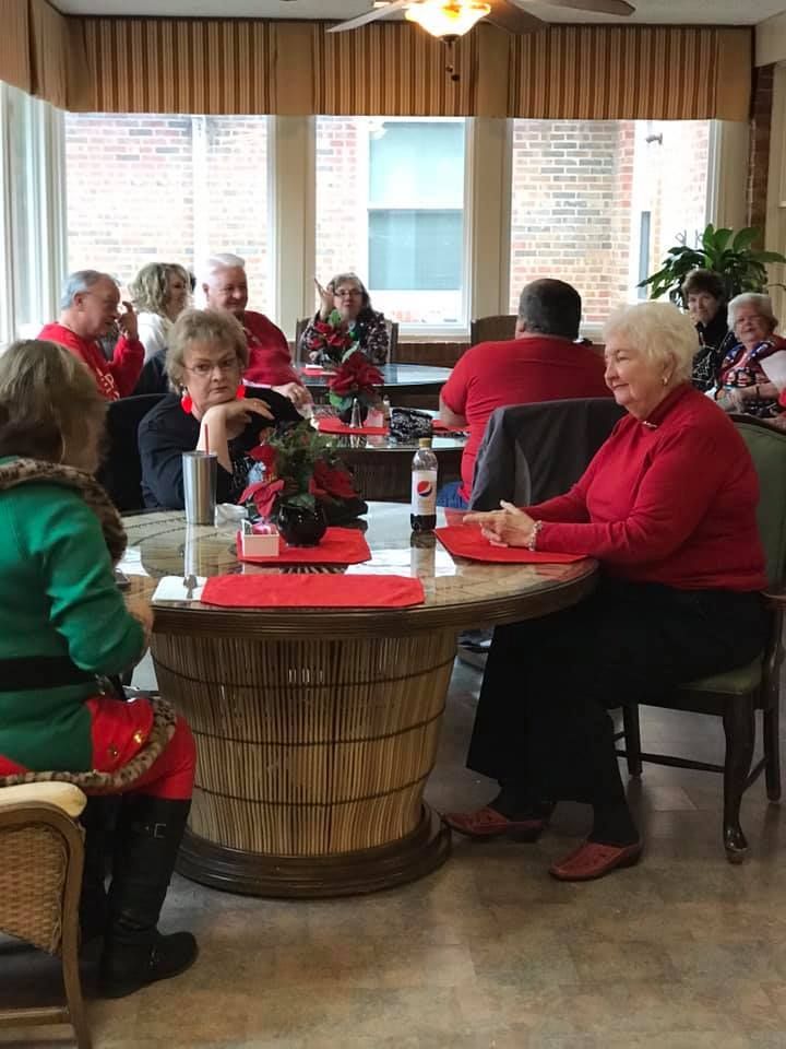 People in a room seated at round tables. Some wear red tops. Christmas decorations are present.