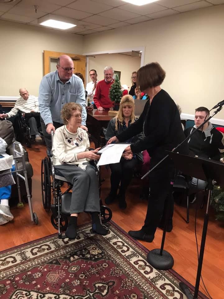 Woman in wheelchair receives document from another woman; room with others.