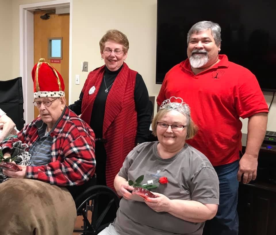 Four people pose for a photo. Two wear crowns and hold roses, one in a wheelchair. All are smiling in a room.