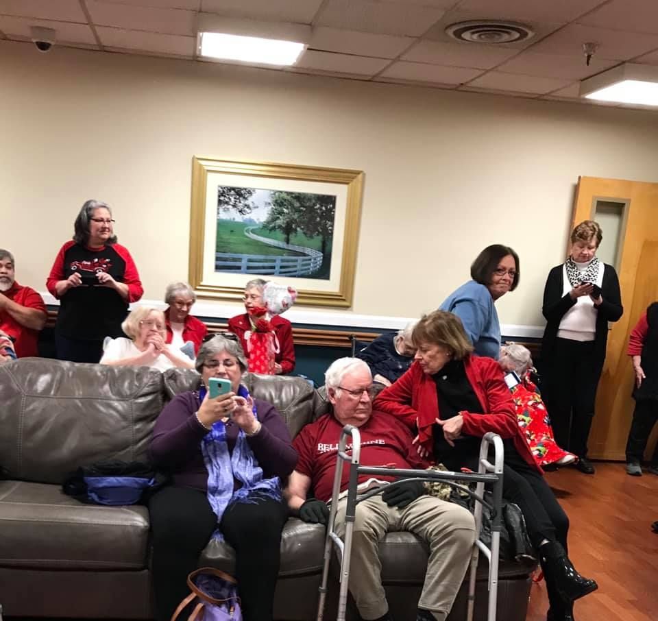 People in a room, some seated, using phones. Some wear red. A framed artwork hangs on a wall.